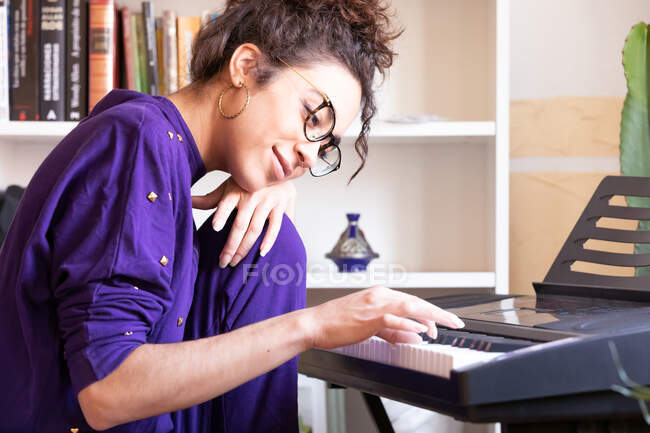 Vue latérale d'une jeune femme hispanique joyeuse jouant du piano électrique tout en pratiquant des compétences musicales à la maison — Photo de stock