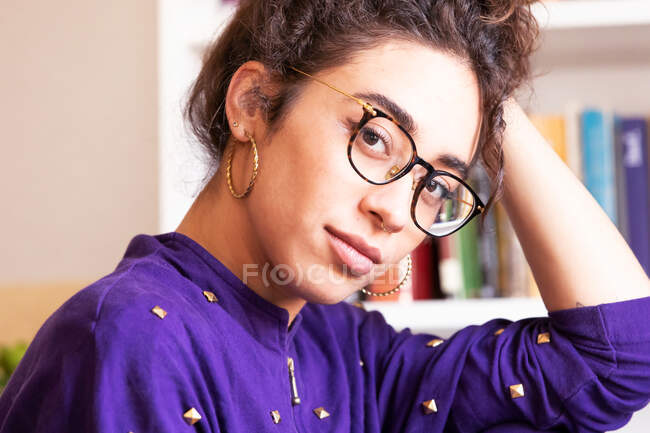 Молодая кудрявая латиноамериканка в повседневной одежде и очках с обручальными сережками, склоняющимися к руке и смотрящими в камеру, стоя возле книжного шкафа дома — стоковое фото