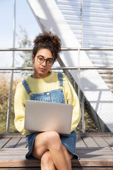 Young Hispanic curly haired woman in denim outfit and glasses looking at camera while sitting with laptop on wooden bench in enclosed urban passage — Stock Photo
