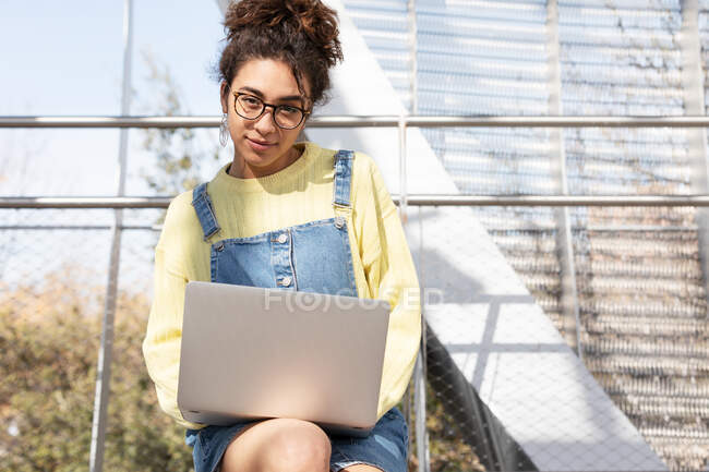 Young Hispanic curly haired woman in denim outfit and glasses looking at camera while sitting with laptop on wooden bench in enclosed urban passage — Stock Photo