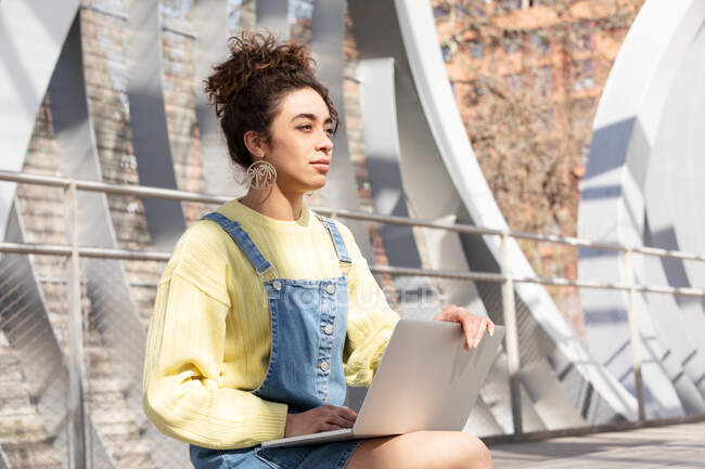 Young Hispanic curly haired woman in denim outfit looking away while sitting with laptop on wooden bench in enclosed urban passage — Stock Photo