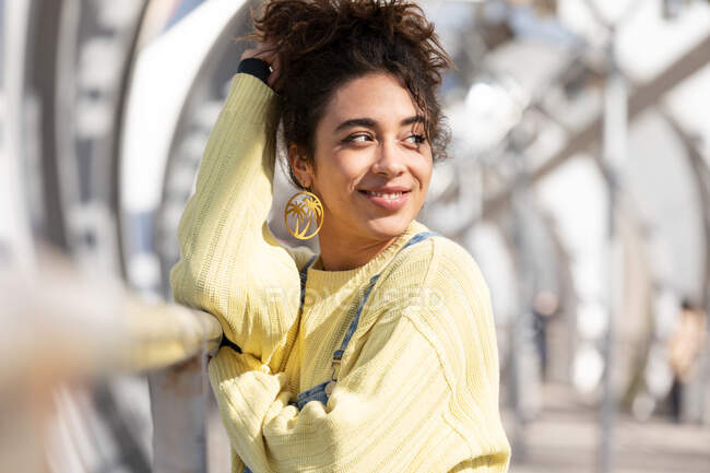 Confident Hispanic female teenager with curly hair wearing denim overalls and yellow sweatshirt with earrings looking away while leaning on railing on enclosed urban bridge — Stock Photo
