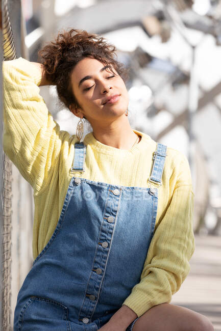 Adolescente hispânica confiante com cabelo encaracolado vestindo macacão jeans e camisola amarela com brincos sentados com os olhos fechados na ponte urbana fechada — Fotografia de Stock