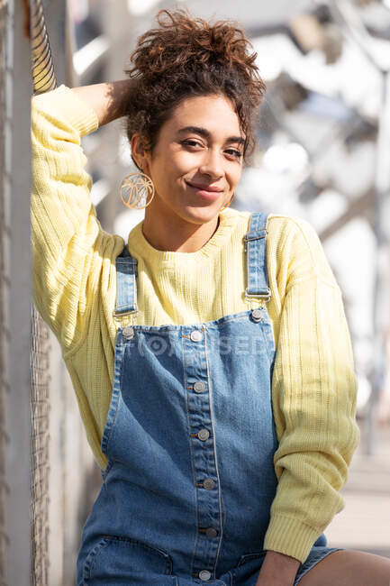 Adolescente hispanique confiante aux cheveux bouclés portant une combinaison en denim et un sweat-shirt jaune avec des boucles d'oreilles regardant la caméra assise sur un pont urbain clos — Photo de stock