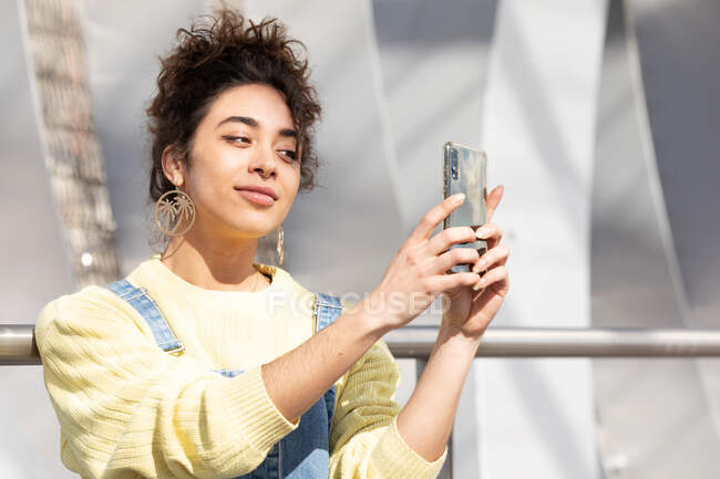 Adolescente hispana sonriente con atuendo informal de moda y pendientes tomando selfie en un teléfono inteligente para compartir en las redes sociales mientras se enfrenta a un entorno urbano borroso - foto de stock