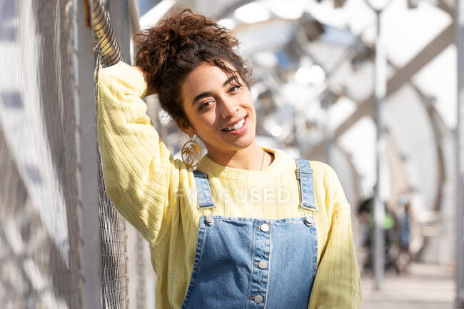 Adolescente hispana confiada con pelo rizado con overoles de mezclilla y sudadera amarilla con pendientes mirando a la cámara mientras está sentada en un puente urbano cerrado - foto de stock