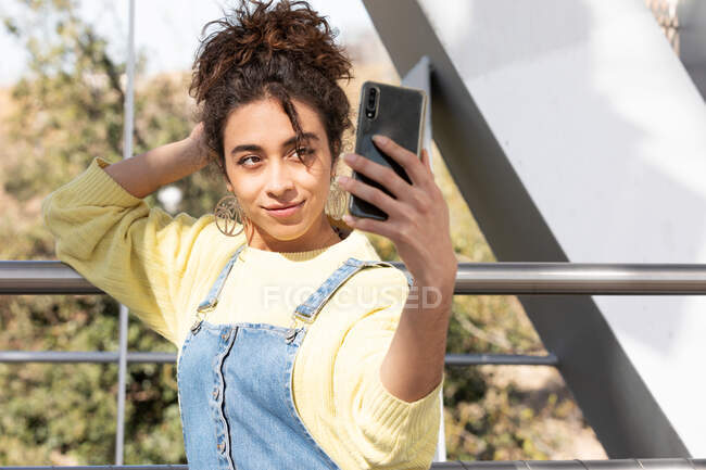 Adolescente hispana sonriente con atuendo informal de moda y pendientes tomando selfie en un teléfono inteligente para compartir en las redes sociales mientras se enfrenta a un entorno urbano borroso - foto de stock
