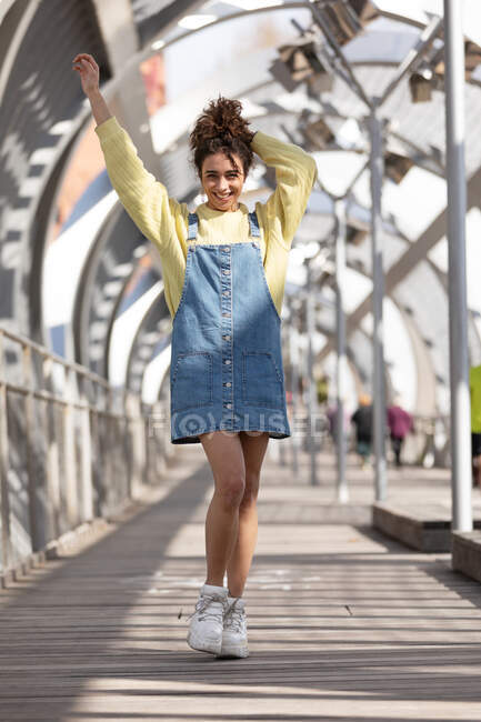 Low angle full body of happy young Hispanic female with curly hair wearing denim overall dress with yellow sweatshirt and sneakers walking on enclosed footbridge in city — Stock Photo