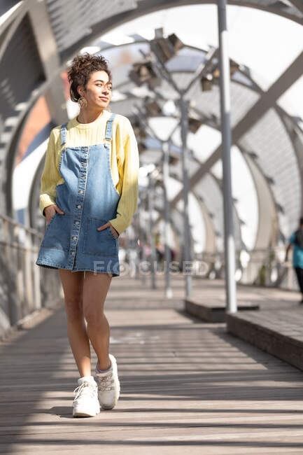 Full body of happy young Hispanic female with curly hair wearing denim overall dress with yellow sweatshirt and sneakers walking looking away on enclosed footbridge in city — Stock Photo