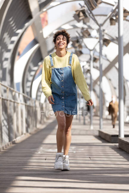 Low angle full body of happy young Hispanic female with curly hair wearing denim overall dress with yellow sweatshirt and sneakers walking on enclosed footbridge in city — Stock Photo