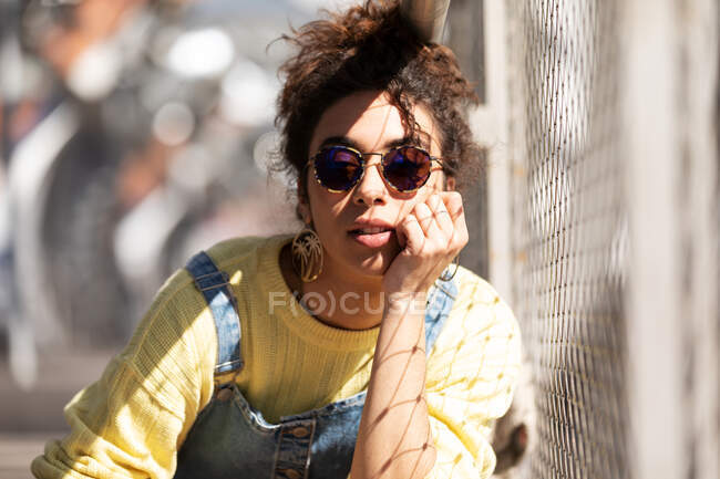 Modern millennial Hispanic female with curly hair wearing yellow sweatshirt with denim overalls and trendy sunglasses and earrings sitting leaning on hand near mesh fence in sunlight — Stock Photo