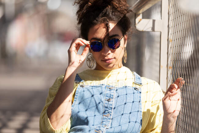 Moderna mujer hispana milenaria con el pelo rizado usando sudadera amarilla con overoles de mezclilla y gafas de sol de moda y pendientes de pie mirando a la cámara cerca de la valla de malla a la luz del sol - foto de stock