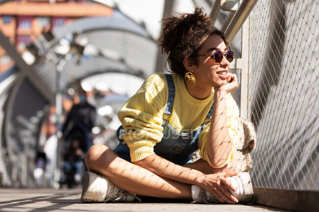 Moderna mujer hispana milenaria con el pelo rizado usando sudadera amarilla con overoles de mezclilla y gafas de sol de moda y pendientes sentados apoyados en la mano mirando hacia otro lado cerca de la valla de malla a la luz del sol - foto de stock