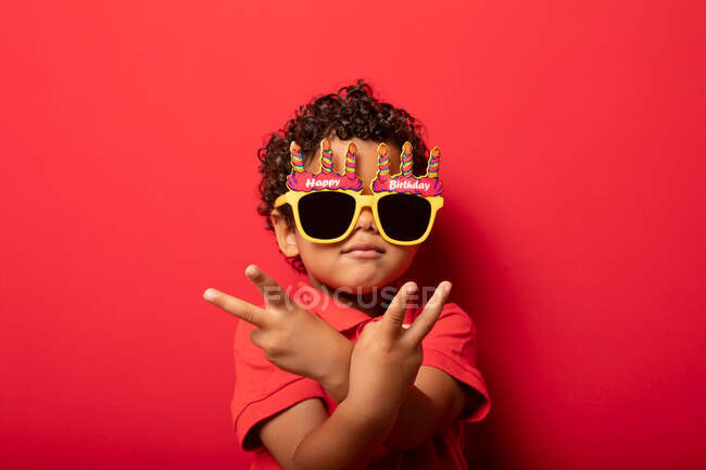 Крутой ребенок в ярких солнечных очках с днем рождения, показывающих мирный жест на красном фоне в студии — стоковое фото