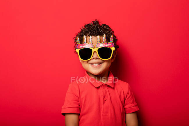 Criança legal vestindo brilhantes óculos de sol Feliz Aniversário no fundo vermelho em estúdio — Fotografia de Stock
