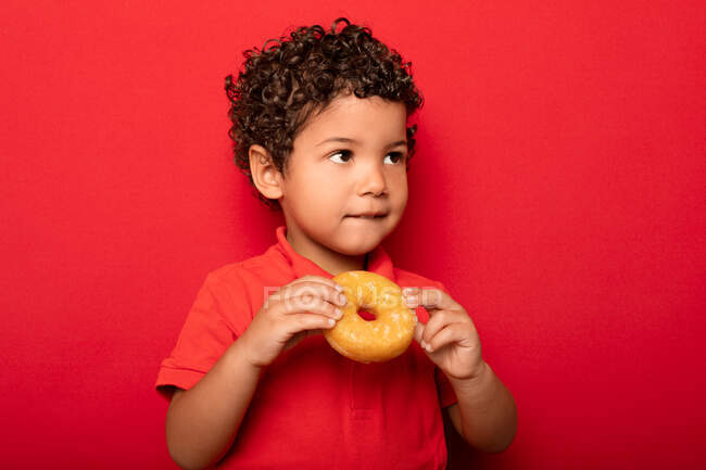 Adorable enfant aux cheveux bouclés mangeant un beignet doux et savoureux et regardant loin sur fond rouge — Photo de stock