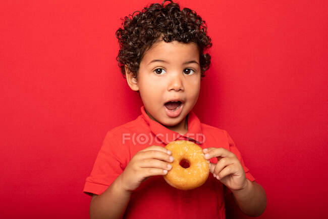 Очаровательный ребенок с вьющимися волосами ест сладкий вкусный пончик и смотрит на камеру на красном фоне — стоковое фото