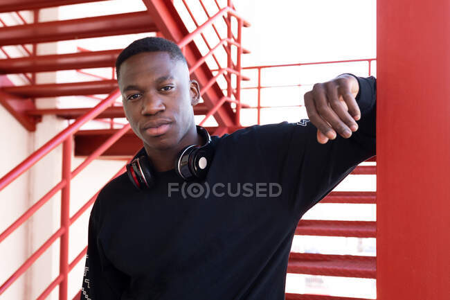 Junger afroamerikanischer Mann mit Kopfhörern am Hals blickt in die Kamera, während er auf einer Metalltreppe steht — Stockfoto