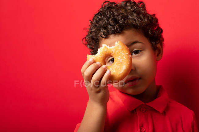 Boy looking at camera through hole in sweet doughnut on red background in studio — Stock Photo
