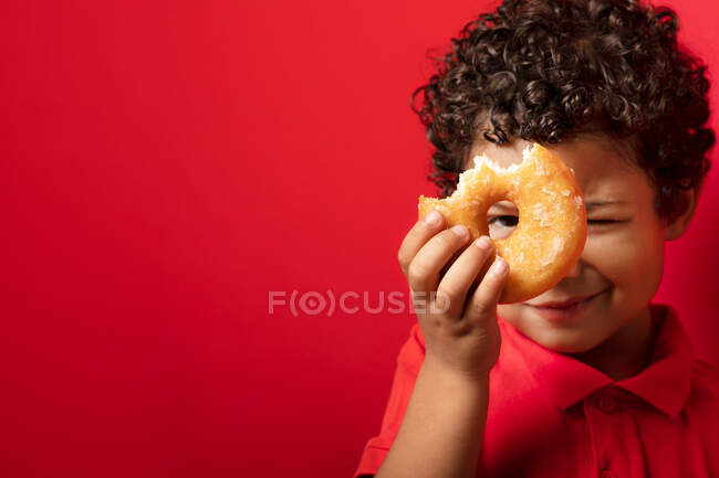 Усміхнений хлопчик дивиться на камеру через отвір у солодкому пончику на червоному тлі в студії — стокове фото