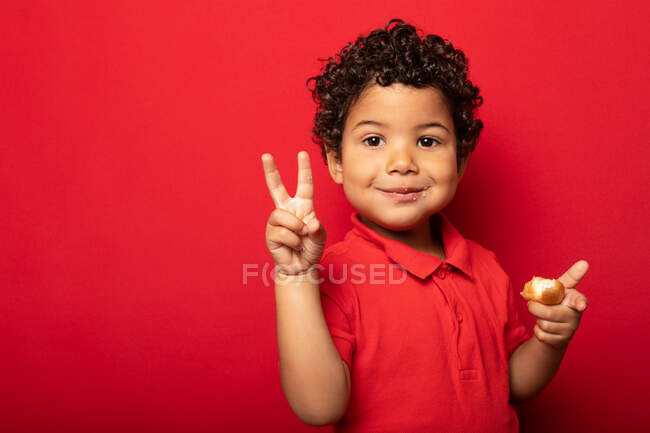 Entzückendes Kind isst köstlichen Donut und zeigt V-Zeichen, während es in die Kamera auf rotem Hintergrund im Studio schaut — Stockfoto