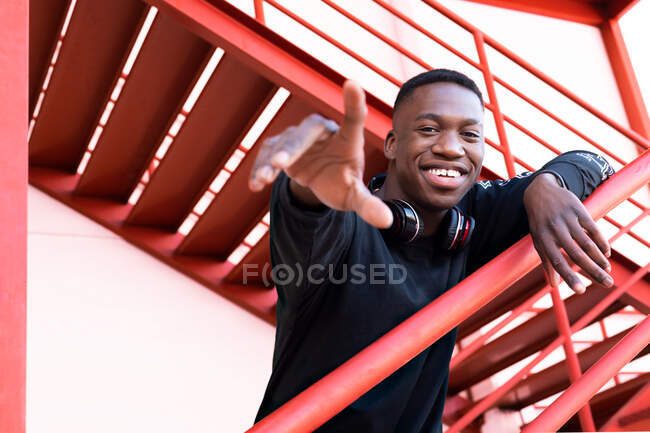 Delighted young African American male with headphones on neck raising arm trying to reach gesture and looking at camera while standing on outdoor metal staircase — Stock Photo