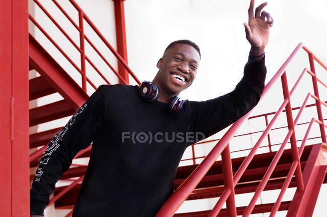 Delighted young African American male with headphones on neck raising arm in greeting gesture and looking at camera while standing on outdoor metal staircase — Stock Photo