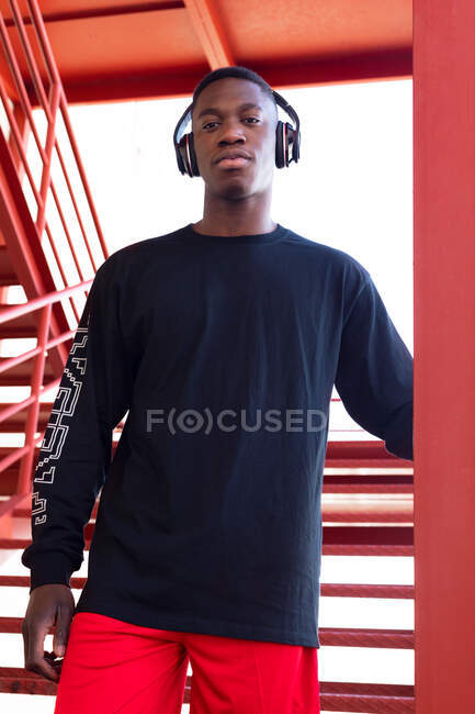 Unemotional African American male in activewear and wireless headphones standing on staircase and looking at camera confidently — Stock Photo