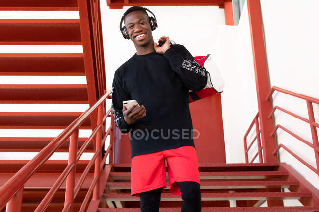Joyeux homme afro-américain en vêtements de sport et écouteurs navigation téléphone mobile et debout sur un escalier en métal avec sac de gym regardant la caméra — Photo de stock