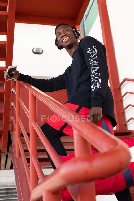 From below joyful African American male in activewear and headphones holding mobile phone and standing on metal staircase with gym bag looking away — Stock Photo