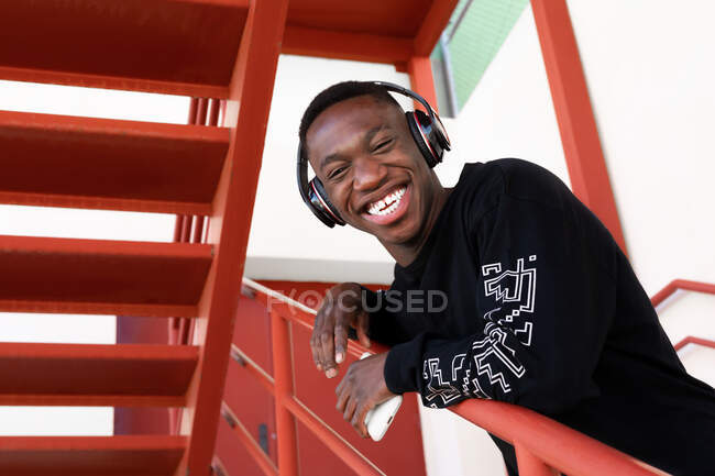 Macho afro-americano alegre em desgaste casual e fones de ouvido sem fio sorrindo olhando para a câmera enquanto estava em pé na escada de metal — Fotografia de Stock