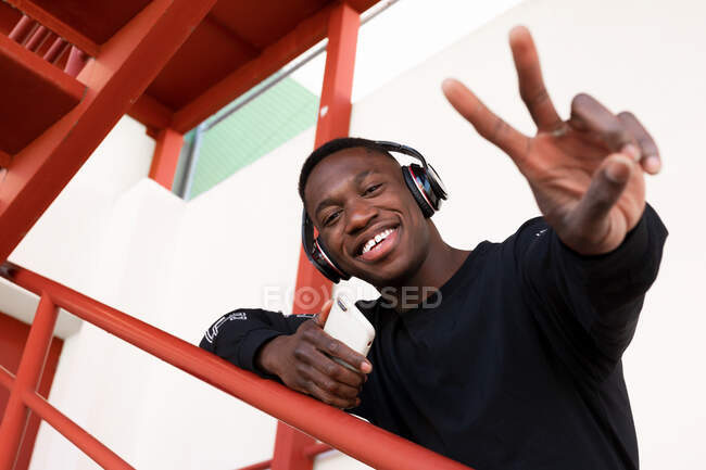Varón afroamericano sonriente de bajo ángulo en ropa casual y auriculares inalámbricos que muestran dos dedos y miran a la cámara mientras está de pie en la escalera de metal - foto de stock