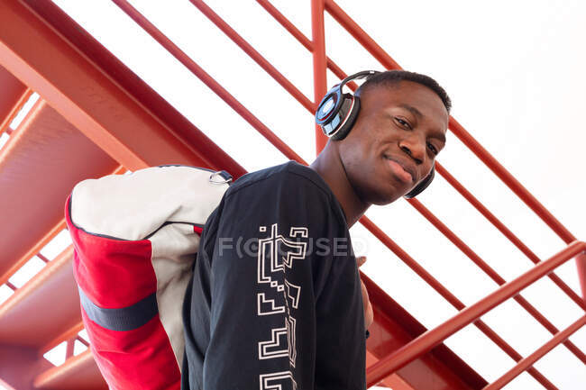 Von unten zufrieden Afroamerikaner in Activwear und kabellosen Kopfhörern, die auf der Treppe stehen und selbstbewusst mit Turntasche in die Kamera schauen — Stockfoto