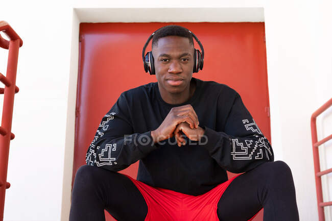 Zufriedener Afroamerikaner in Activwear und drahtlosen Kopfhörern sitzt auf der Treppe und blickt selbstbewusst in die Kamera — Stockfoto