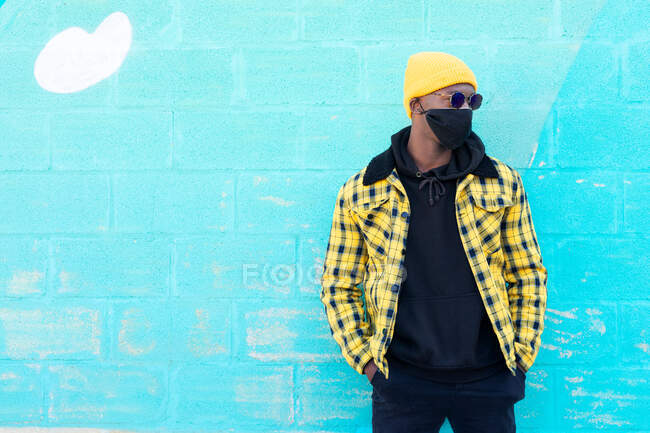 Крутой афроамериканец в модной неформальной одежде, маске для лица и солнцезащитных очках, отводящий взгляд, стоя на улице — стоковое фото