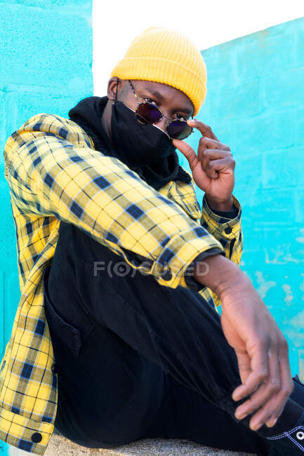 Cool African American male in trendy informal wear and face mask lowering sunglasses and looking at camera while sitting on street — Stock Photo