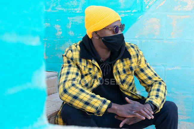 Cool African American male in trendy informal wear and face mask and sunglasses and looking away while sitting on stairs in the street — Stock Photo