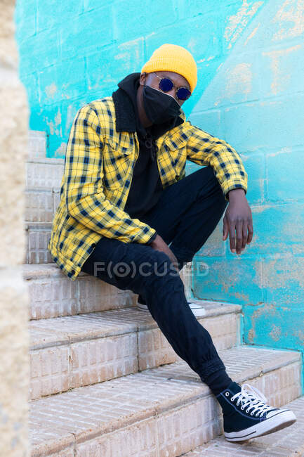 Cool Afro-Américain mâle en tenue informelle tendance et masque facial et lunettes de soleil et regardant la caméra tout en étant assis sur les escaliers dans la rue — Photo de stock