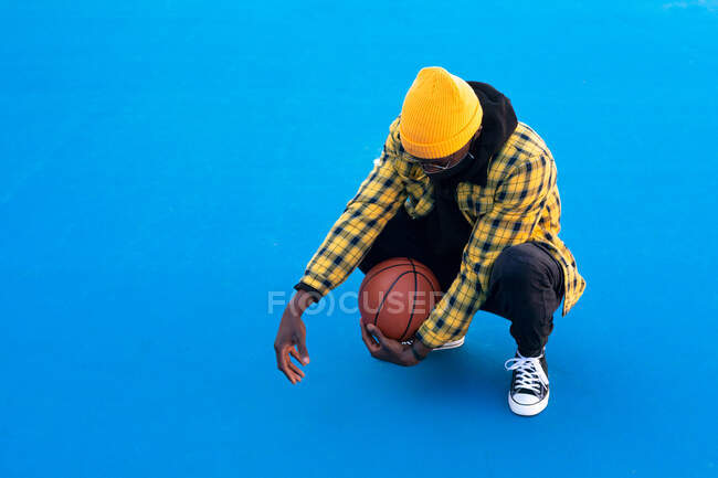 Full length confident African American male in trendy wear and face mask sitting on haunches with basketball ball on court and looking down — Stock Photo