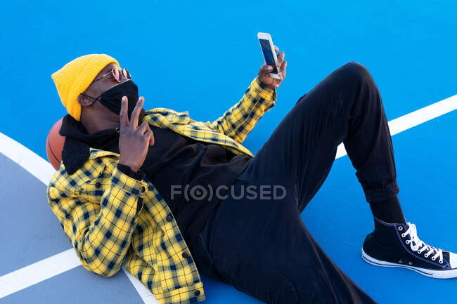 Сверху стильный афроамериканец в маске лежит на баскетбольной площадке и делает селфи на смартфоне, показывая V знак — стоковое фото