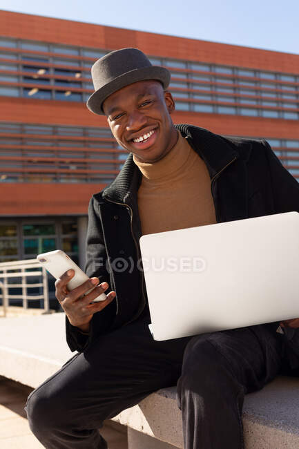 Веселий афроамериканець у стильному одязі і капелюсі, який дивиться мобільний телефон, сидячи на вулиці з нетбуком на колінах, дивлячись на камеру. — стокове фото