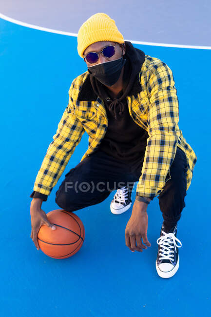 Уверенный в себе афроамериканец в модной одежде и маске для лица сидит на ягодицах с баскетбольным мячом на площадке и смотрит в камеру — стоковое фото