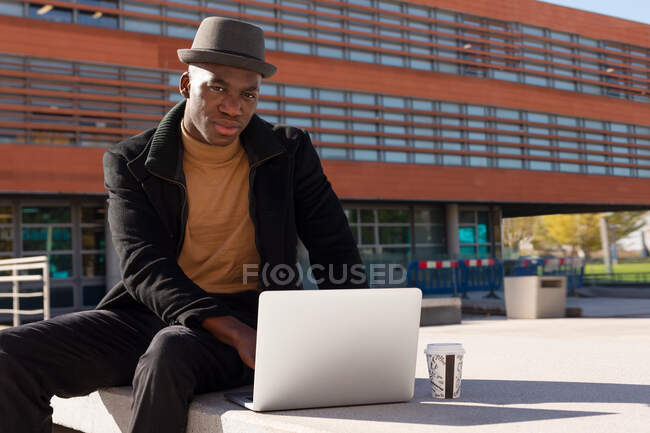 Pensivo afro-americano masculino em roupa elegante usando netbook e com takeaway café sentado na rua ensolarada olhando para a câmera — Fotografia de Stock