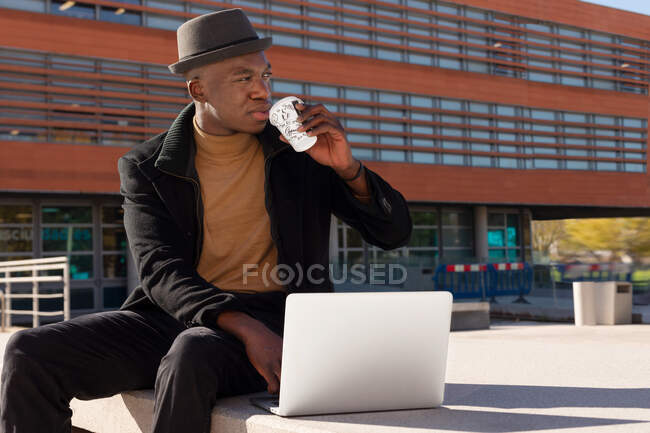 Hombre afroamericano pensativo en traje elegante usando netbook y beber café para llevar mientras está sentado en la calle soleada - foto de stock