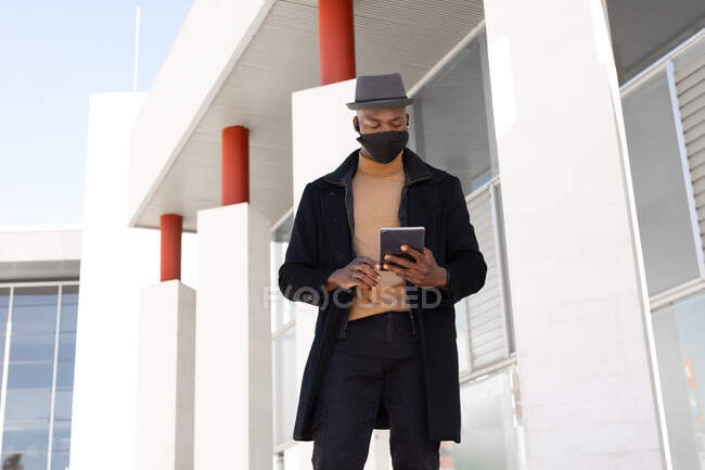 Faible angle positif Afro-Américain mâle dans l'usure élégante et masque de navigation tablette tout en se tenant debout sur la rue ensoleillée — Photo de stock