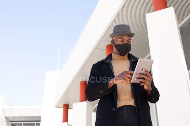 Низкий угол положительный афроамериканец в стильной одежде и маске для лица просмотра планшета, стоя на солнечной улице — стоковое фото