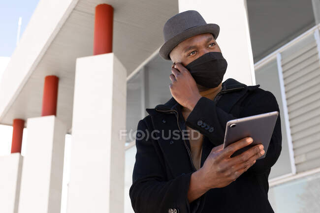 Bajo ángulo positivo afroamericano masculino en elegante desgaste y máscara de la cara tableta de navegación mientras está de pie en la calle soleada mirando hacia otro lado - foto de stock