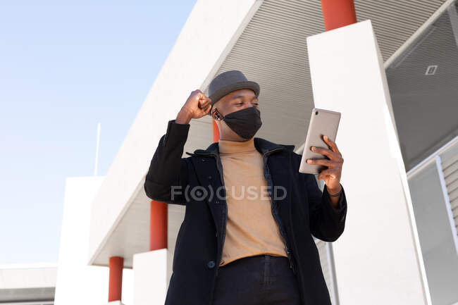 Low angle positive African American male in stylish wear and face mask doing video call on tablet while with clenched fist celebrating victory with gesture standing on sunny street — Stock Photo