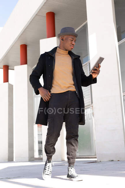 Fröhlicher afroamerikanischer Mann in stylischer Kleidung und Ohrhörern stöbert auf der sonnigen Straße in modernen Tablets und schaut lächelnd auf den Bildschirm — Stockfoto