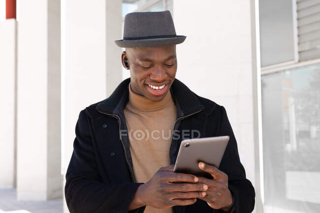 Homme afro-américain joyeux dans des vêtements élégants et écouteurs naviguant tablette moderne sur la rue ensoleillée et regardant l'écran avec le sourire — Photo de stock